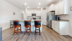 A modern kitchen with white cabinets, a central island with bar stools, stainless steel appliances, and a "JOY" sign on the wall. - Lovely coastal home with pool access, fast WiFi, screened patio & great location (Port St. Joe)