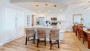 A modern kitchen with white cabinets, a central island, and three wicker chairs. - Renovated oceanfront condo with lanai, pool, hot tub & view - snowbird-friendly (Mexico Beach)
