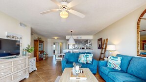 A spacious living room with a blue sofa, a wooden coffee table, and a television. - Gulf-front condo with balcony, ocean view, W&D, pool, & fitness room (Fort Walton Beach)