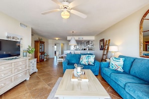 A spacious living room with a blue sofa, a wooden coffee table, and a television. - Gulf-front condo with balcony, ocean view, W&D, pool, & fitness room (Fort Walton Beach)