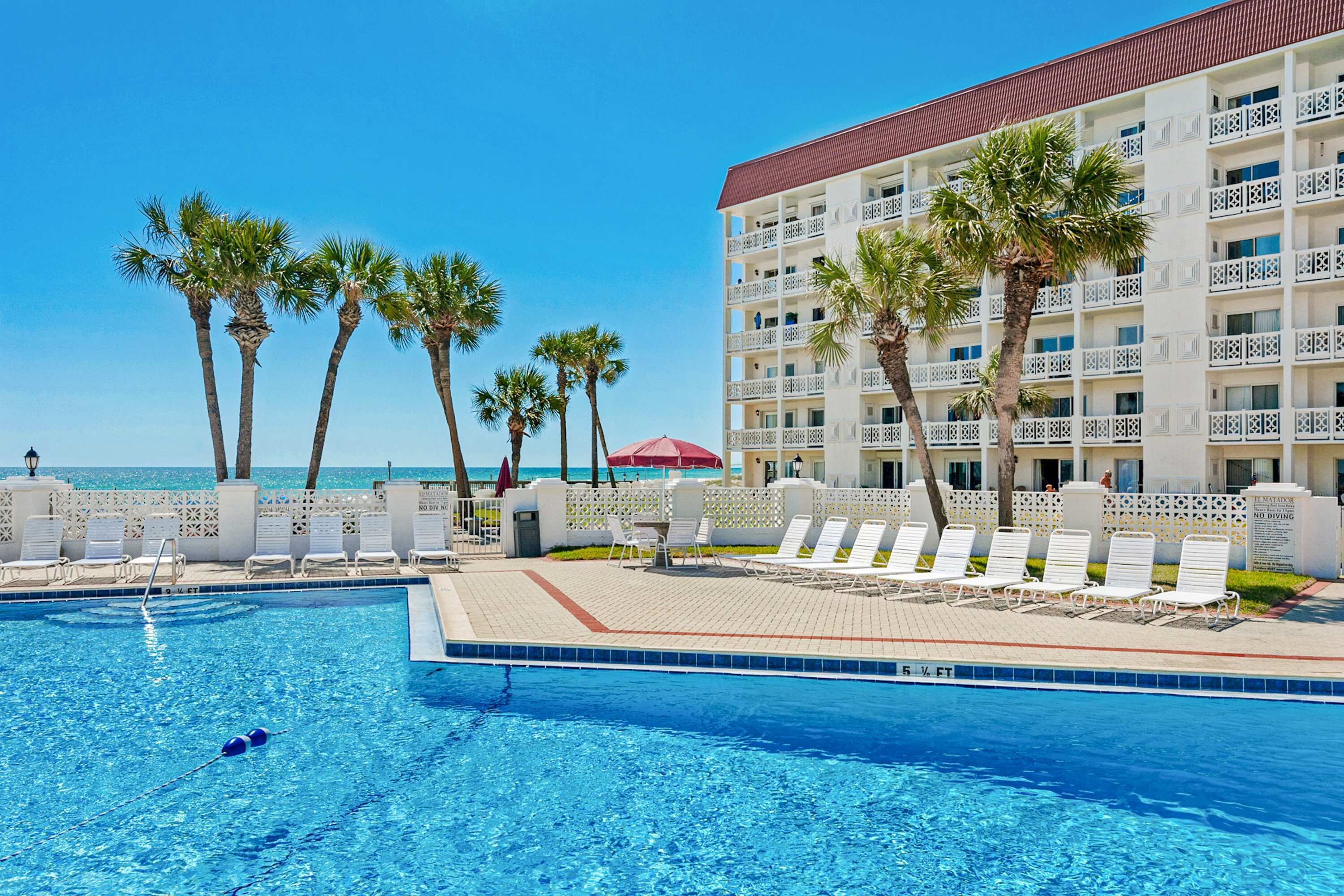 A pool area with lounge chairs, palm trees, and a building with balconies.