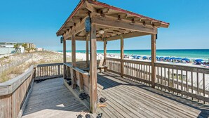 Eine Holzstege mit überdachter Sitzgelegenheit mit Blick auf einen Strand mit Sonnenschirmen und Menschen.