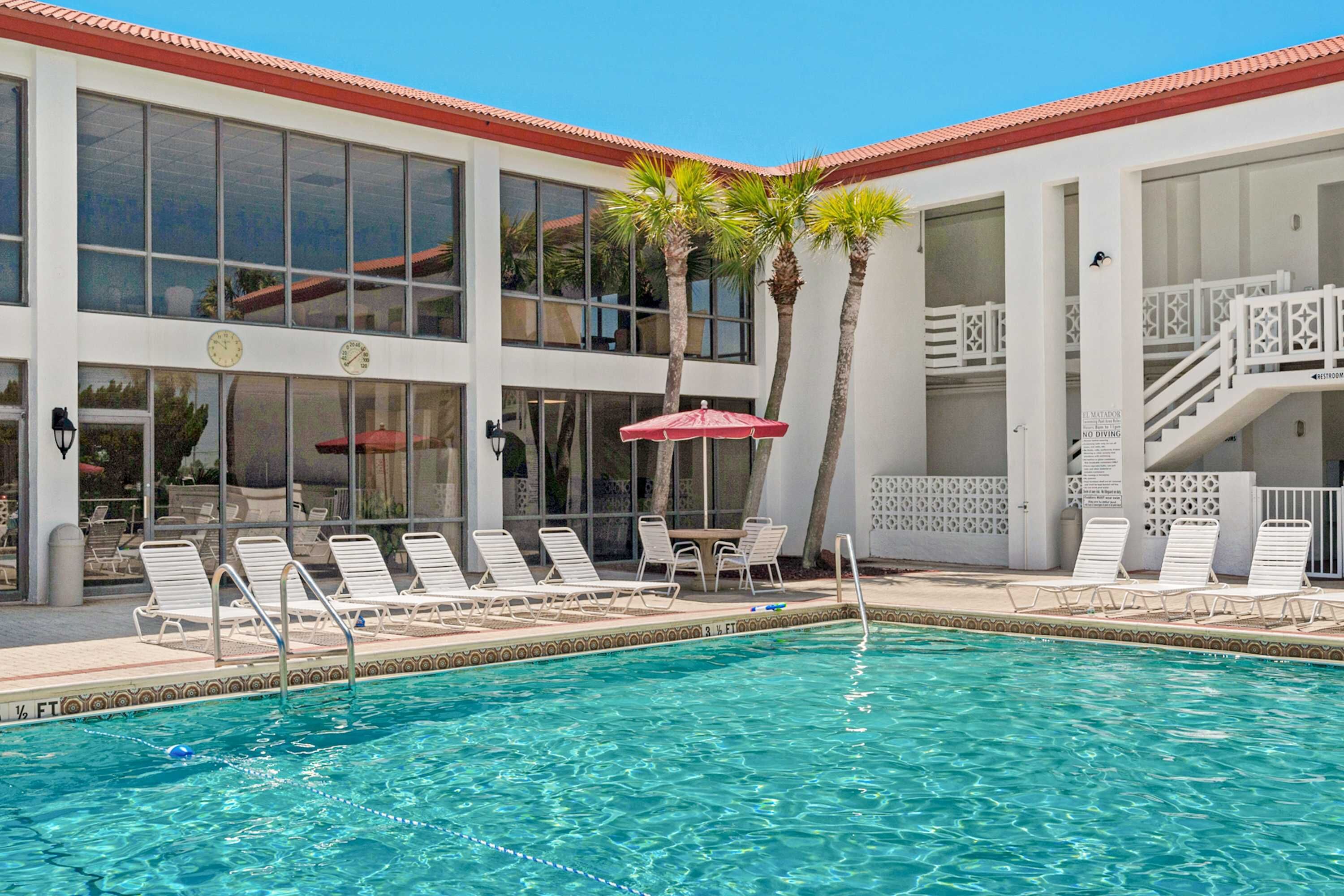 A pool area with lounge chairs, palm trees, and a building with large windows and a red roof.