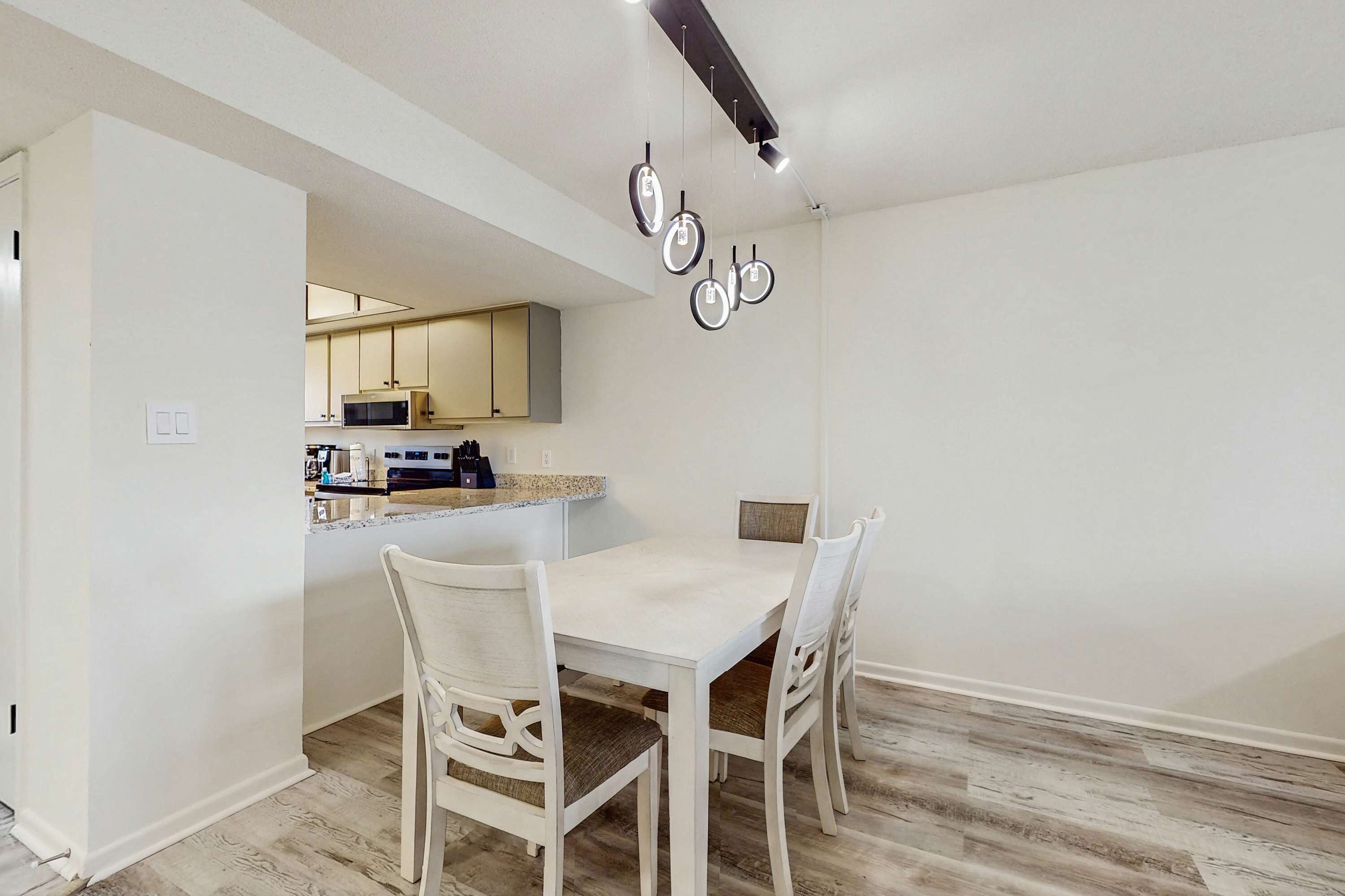 A dining area with a white table and chairs, a kitchen in the background, and a ceiling light fixture.