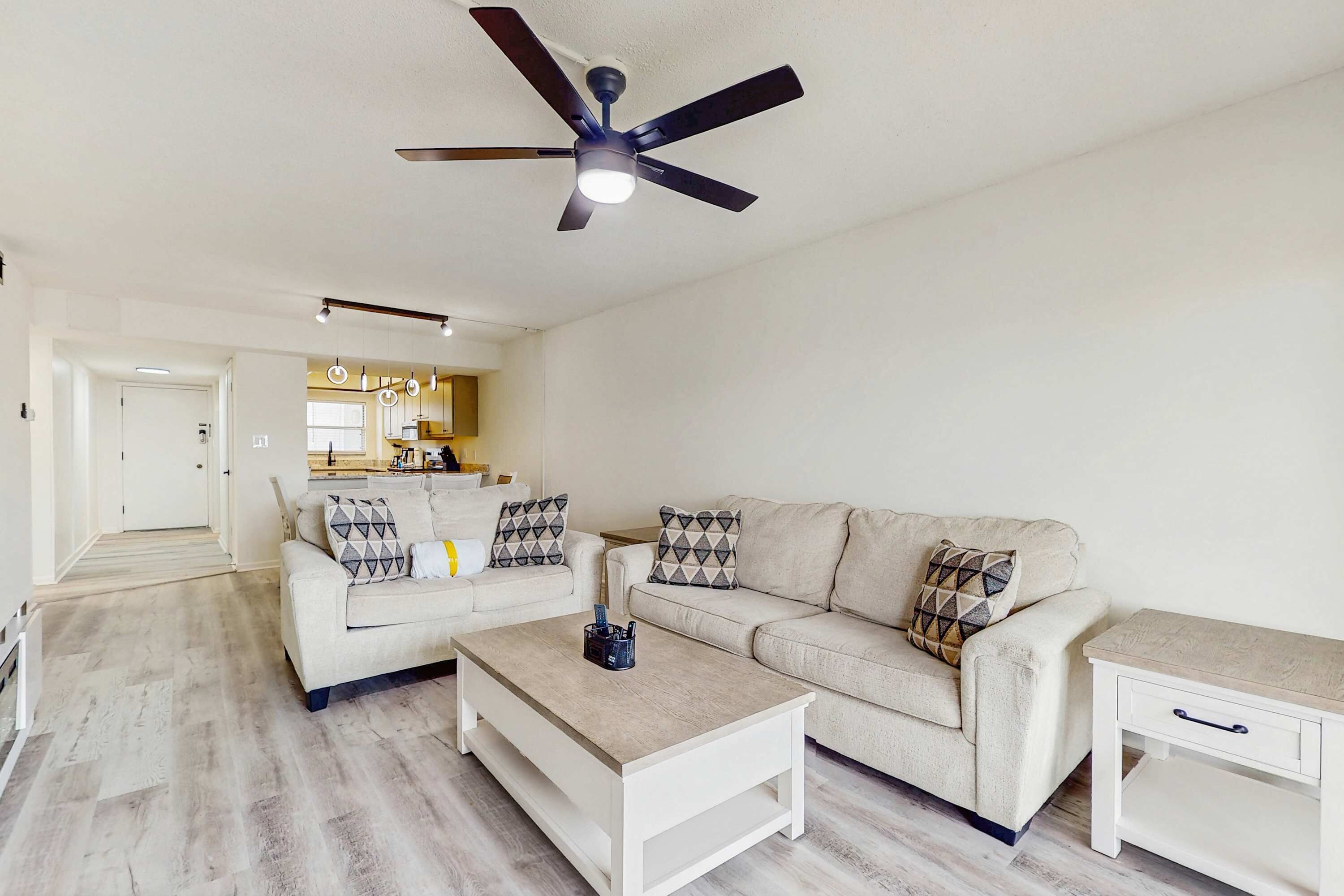 Ein Wohnzimmer mit einem beigen Sofa, einem weißen Couchtisch und einem kleinen weißen Beistelltisch. Über der Möbeln ist ein Deckenventilator zu sehen.