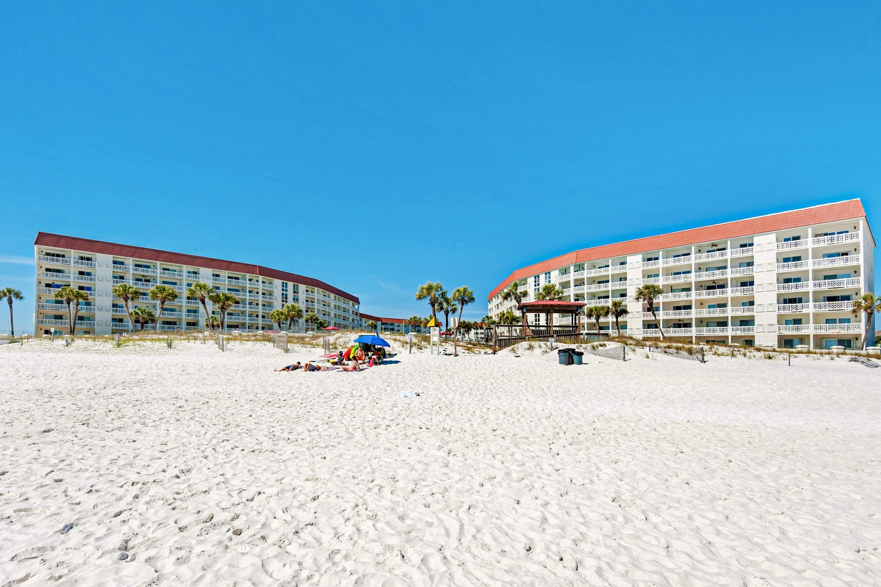 Ein Strandhotel mit mehreren Etagen, Palmen und einem Sandstrand im Vordergrund.