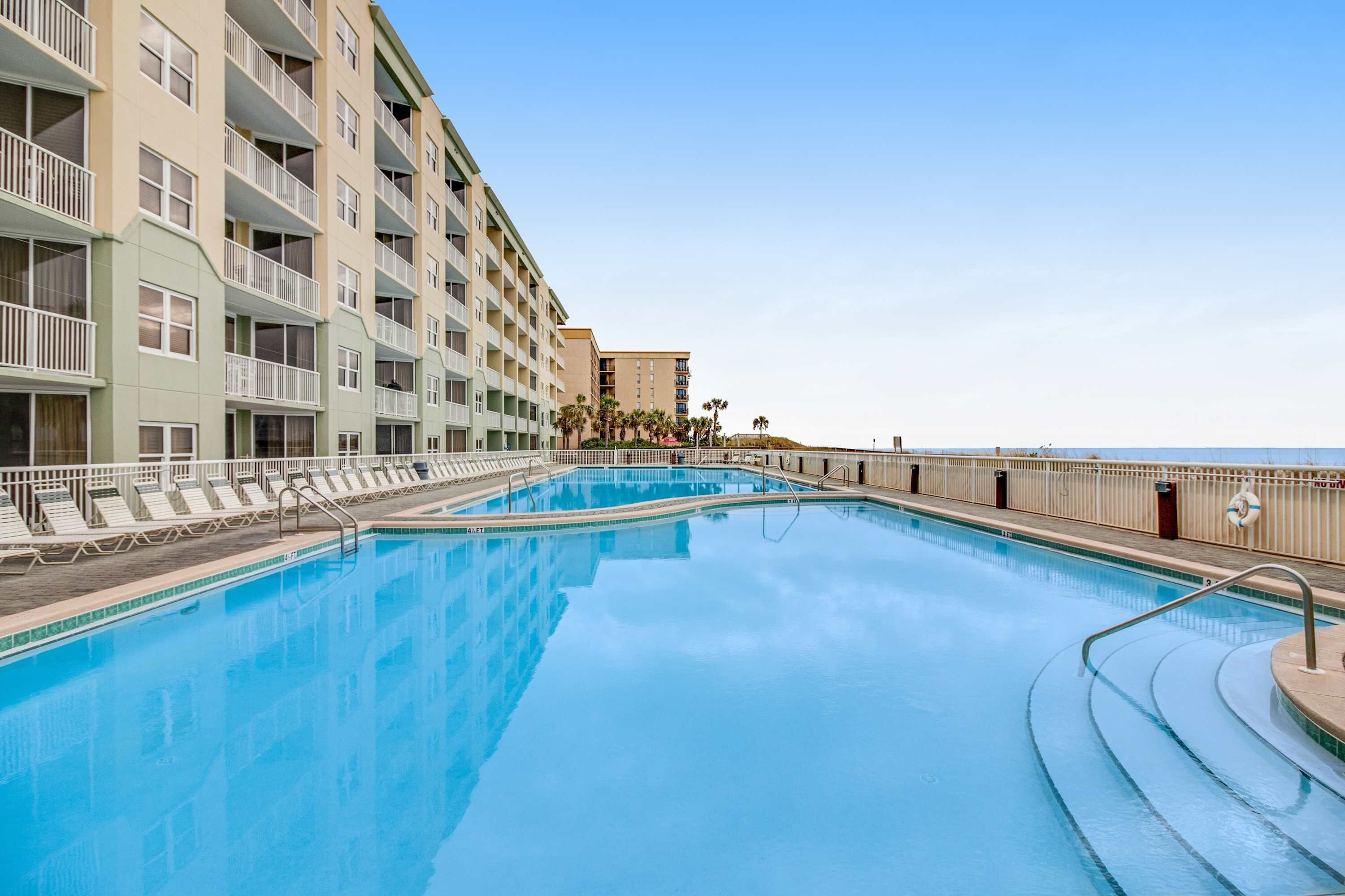A swimming pool adjacent to a multi-story hotel with balconies, a lifeguard stand, and a clear view of the ocean.