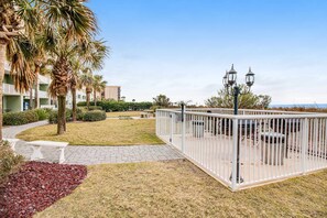 A courtyard with a white fence, benches, and a pathway leading to a building. - Gulf front condo with furnished patio, pool, fitness room, boardwalk to beach (Fort Walton Beach)