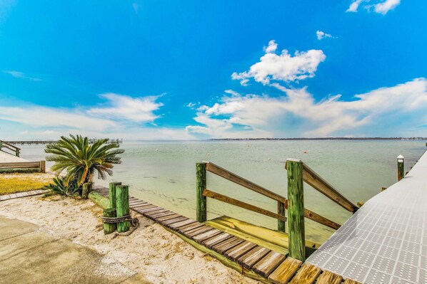 A wooden pier leading to a sandy beach with clear water and a palm tree. - Stunning coastal retreat featuring roomy interiors and quick beach access (Pensacola Beach)