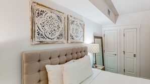 Een slaapkamer met een gestoffeerd hoofdeinde, witte beddengoed, twee decoratieve wanddecoraties, een bedlamp en een kledingkast.