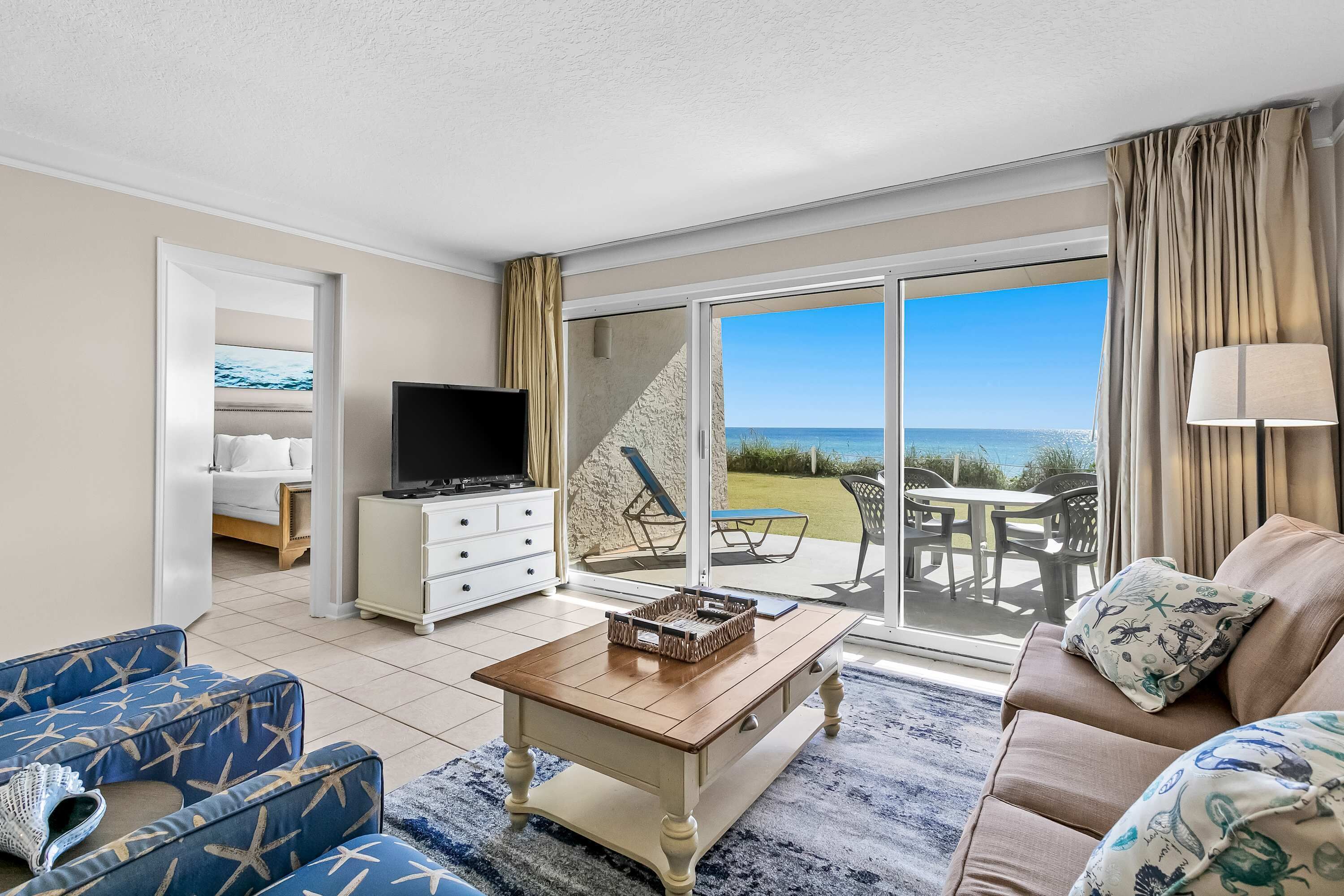 Ein geräumiges Wohnzimmer mit Fernseher, Couchtisch und Sofa. Eine Glasschiebetür führt zu einer Terrassenanlage mit Meerblick.