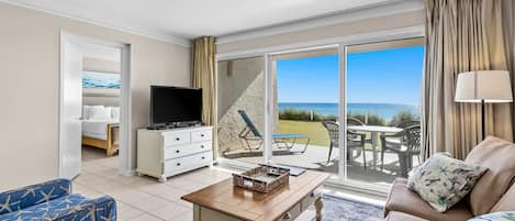 A spacious living room with a television, a coffee table, and a sofa. A sliding glass door leads to an outdoor patio with a view of the ocean.