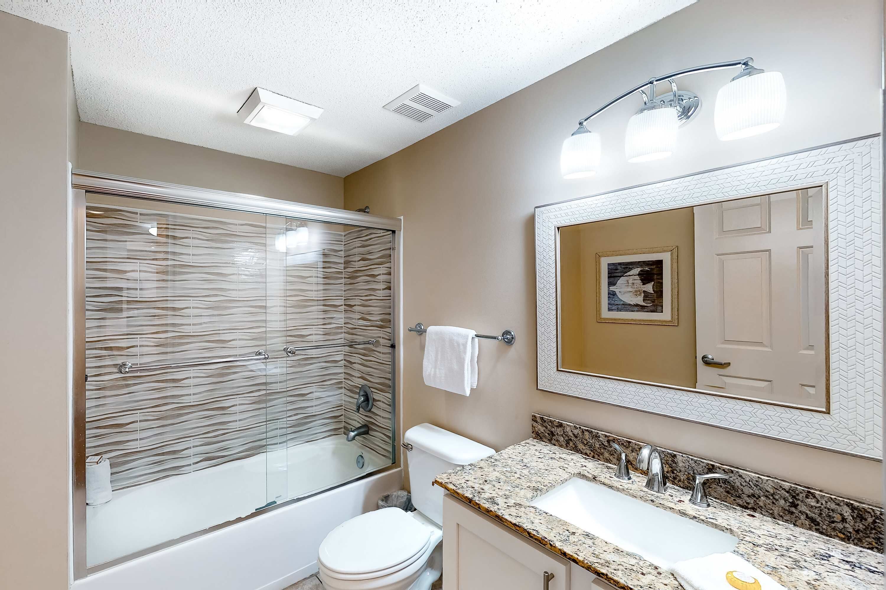 Une salle de bain moderne dotée d’une douche à parois de verre, de toilettes blanches, d’un lavabo avec un plan de travail en granit et d’un miroir orné d’un cadre décoratif.