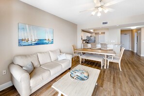 A living room with a beige sofa, a coffee table, and a painting of sailboats. - 2nd Floor Coastal Condo On Okaloosa Island Gulf-Front Views (Fort Walton Beach)
