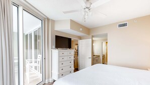 Habitación de hotel con una cama grande, ventilador de techo, televisión, puerta corrediza de vidrio que da a un balcón y una kitchenette.