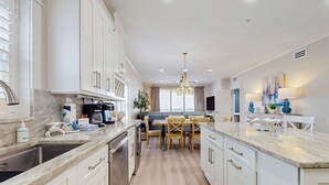 Una cocina moderna con armarios blancos, encimera de mármol y un comedor con sillas y mesa de madera.