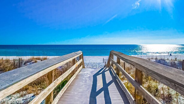 Un paseo marítimo de madera que conduce a una playa con cielos despejados y luz solar.