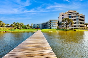 A wooden pier leading to a modern building with multiple floors, surrounded by greenery and a calm body of water. - Peaceful corner studio with pool & Santa Rosa Sound views - snowbird-friendly (Fort Walton Beach)