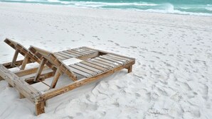 Ein Holzliegestuhl am sandigen Strand, im Hintergrund das Meer mit Wellen.