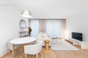 Ein modernes Wohnzimmer mit einem runden Esstisch, einer Couch, einem Fernseher und einem großen Fenster mit transparenten Vorhängen.