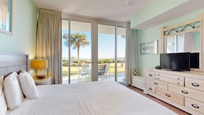 A hotel room with a large bed, a TV, a dresser, and a sliding glass door leading to an outdoor area with a palm tree. - Spacious Ground Floor Condo Steps To Beach, Seasonal Beach Chairs & Umbrella (Fort Walton Beach)