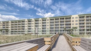 A wooden boardwalk leading to a multi-story building with balconies, overlooking a beach. - Spacious Ground Floor Condo Steps To Beach, Seasonal Beach Chairs & Umbrella (Fort Walton Beach)