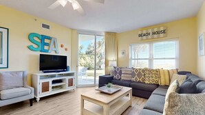 A living room with a TV, a sofa, a coffee table, and a sign that reads "BEACH HOUSE". - Spacious Ground Floor Condo Steps To Beach, Seasonal Beach Chairs & Umbrella (Fort Walton Beach)