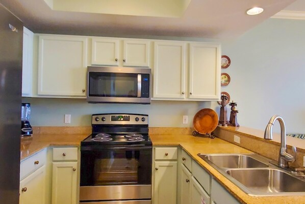 A compact kitchen with white cabinets, a stainless steel microwave, an oven, a sink, and a countertop with a blender. - Beachfront 3rd Floor Condo w Private Balcony, Shared Heated Pool, Beach Access (Fort Walton Beach)
