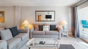 Ein modernes Wohnzimmer mit zwei Sofas, einem Glastisch und einem Flachbildfernseher. Auf Beistelltischen stehen zwei Lampen und ein großes Fenster mit Vorhängen.