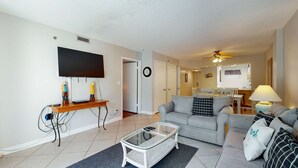 Ein Wohnzimmer mit Flachbildfernseher, Couchtisch und Sofa. Im Hintergrund ist ein Essbereich mit Tisch und Stühlen zu sehen.