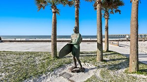 A statue of a person holding a surfboard, surrounded by palm trees and a clear blue sky. - Second-story waterfront condo with pool view, W/D, AC - perfect location (Pensacola Beach)