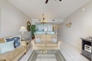 A living room with a beige sofa, a glass coffee table, and a kitchen in the background. - Majestic Sun B305 | Beachfront Bliss! (Miramar Beach)