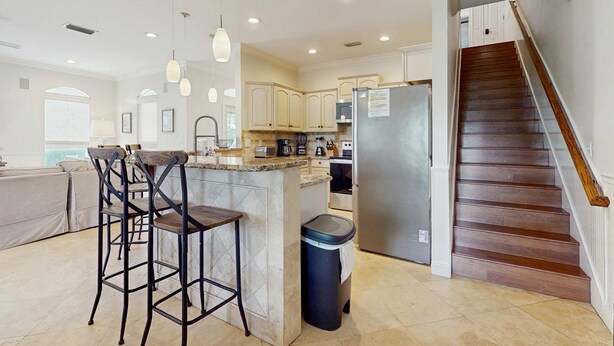 A modern kitchen with a central island, bar stools, a stainless steel refrigerator, and a wooden staircase. - Gorgeous coastal home with plenty of space with shared pool & beach access (Miramar Beach)