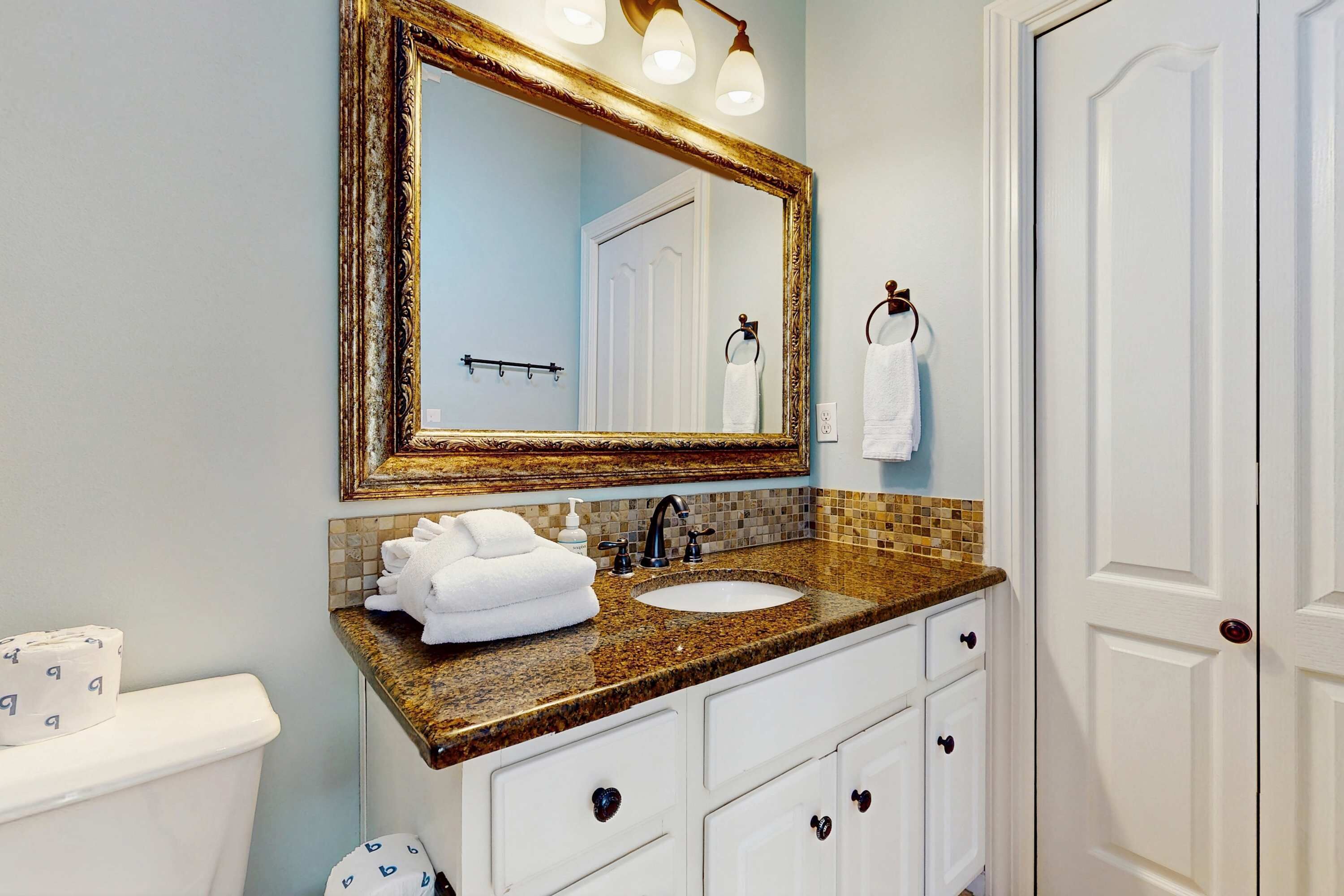A bathroom with a granite countertop, white cabinets, a gold-framed mirror, and a white door.