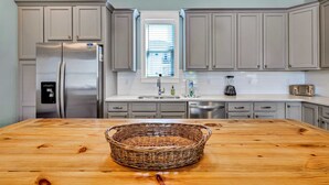 A modern kitchen with grey cabinets, a stainless steel refrigerator, a wooden table, and a window with blinds. - Koko Kabana in Hidden Palms (Miramar Beach)