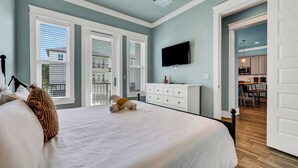 A bedroom with a large bed, a flat-screen TV mounted on the wall, and a view of the kitchen through an open door. - Koko Kabana in Hidden Palms (Miramar Beach)