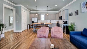 A spacious living area with a wooden floor, a blue sofa, and two pink armchairs. A kitchen with white cabinets and a dining table is visible in the background. - Koko Kabana in Hidden Palms (Miramar Beach)
