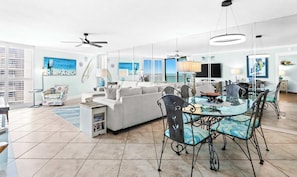 Un vaste espace de vie comprenant un canapé moderne, une table à manger en verre avec des chaises, un ventilateur de plafond et des œuvres d’art accrochées aux murs.