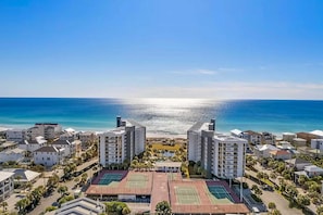 A coastal residential area with multiple high-rise buildings, tennis courts, and a clear view of the ocean. - Gulf-front condo with pool, hot tub & included beach service (Miramar Beach)