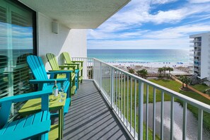A balcony with turquoise chairs and a table, overlooking a beach with a clear view of the ocean and a hotel building in the background. - Gulf-front condo with pool, hot tub & included beach service (Miramar Beach)