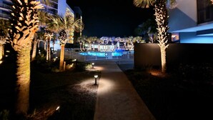 Vista noturna de um resort com piscina, palmeiras e edifícios iluminados.