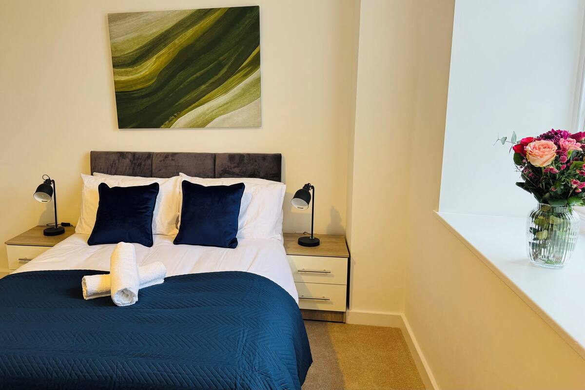 Un lit bien fait, recouvert d’une couverture bleu foncé, avec du linge de lit blanc et deux oreillers bleu foncé. Il y a deux tables de chevet avec des lampes, un vase avec des fleurs sur la droite et un tableau accroché au mur au-dessus du lit.