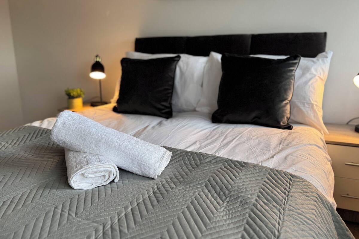 Un lit bien fait, avec une couverture grise, des oreillers blancs et une serviette blanche roulée posée sur le lit.