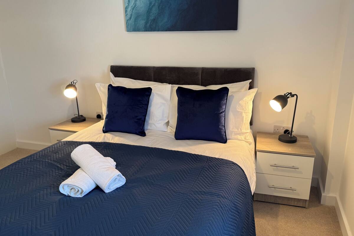 Un lit bien fait, recouvert d’une couverture bleu foncé et agrémenté de deux oreillers blancs, flanqué de deux tables de chevet avec des lampes, dans une pièce simple avec un mur uni et une seule œuvre d’art au-dessus du lit.