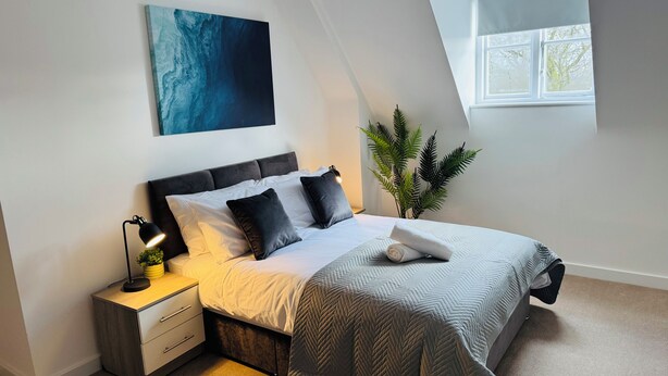 Un dormitorio con cama, mesita de noche, lámpara, planta en maceta y ventana.