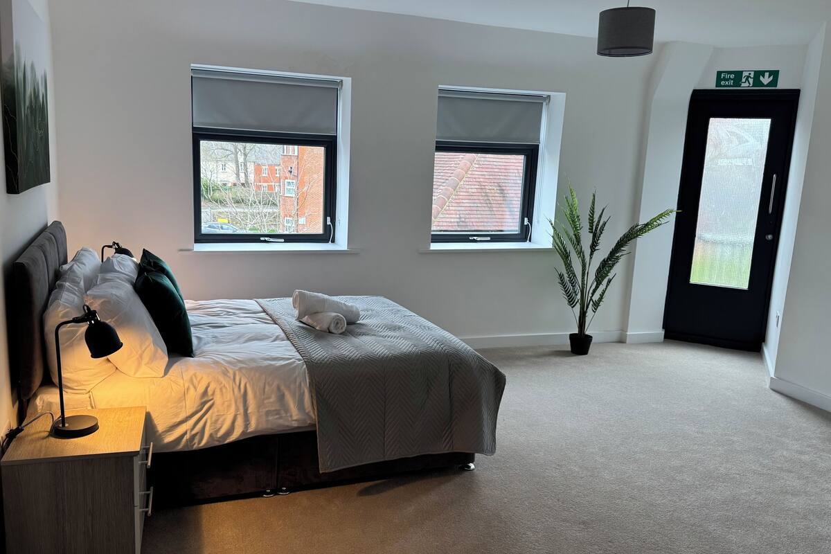 Une chambre à coucher avec un lit, une lampe de chevet, une plante en pot et une vue sur l’extérieur grâce à deux fenêtres.