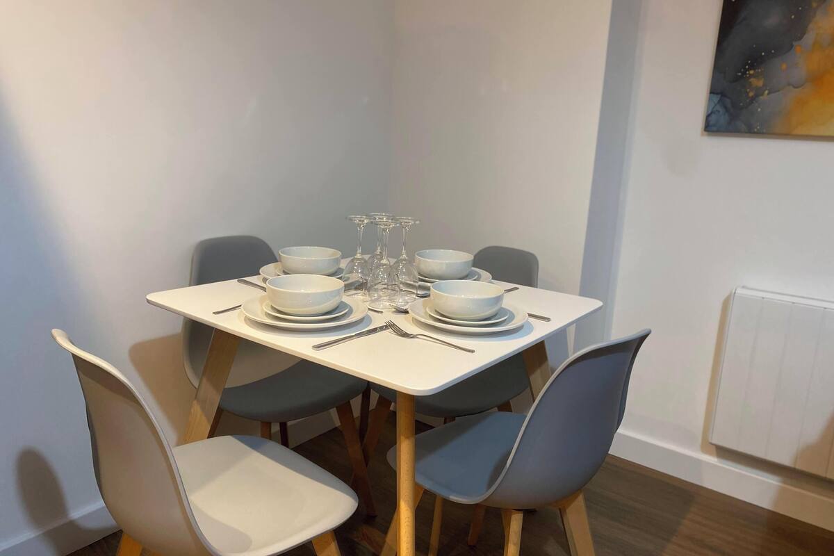 Une table à manger dressée avec de l’assiette, des bols et des couverts blancs, entourée de chaises grises.
