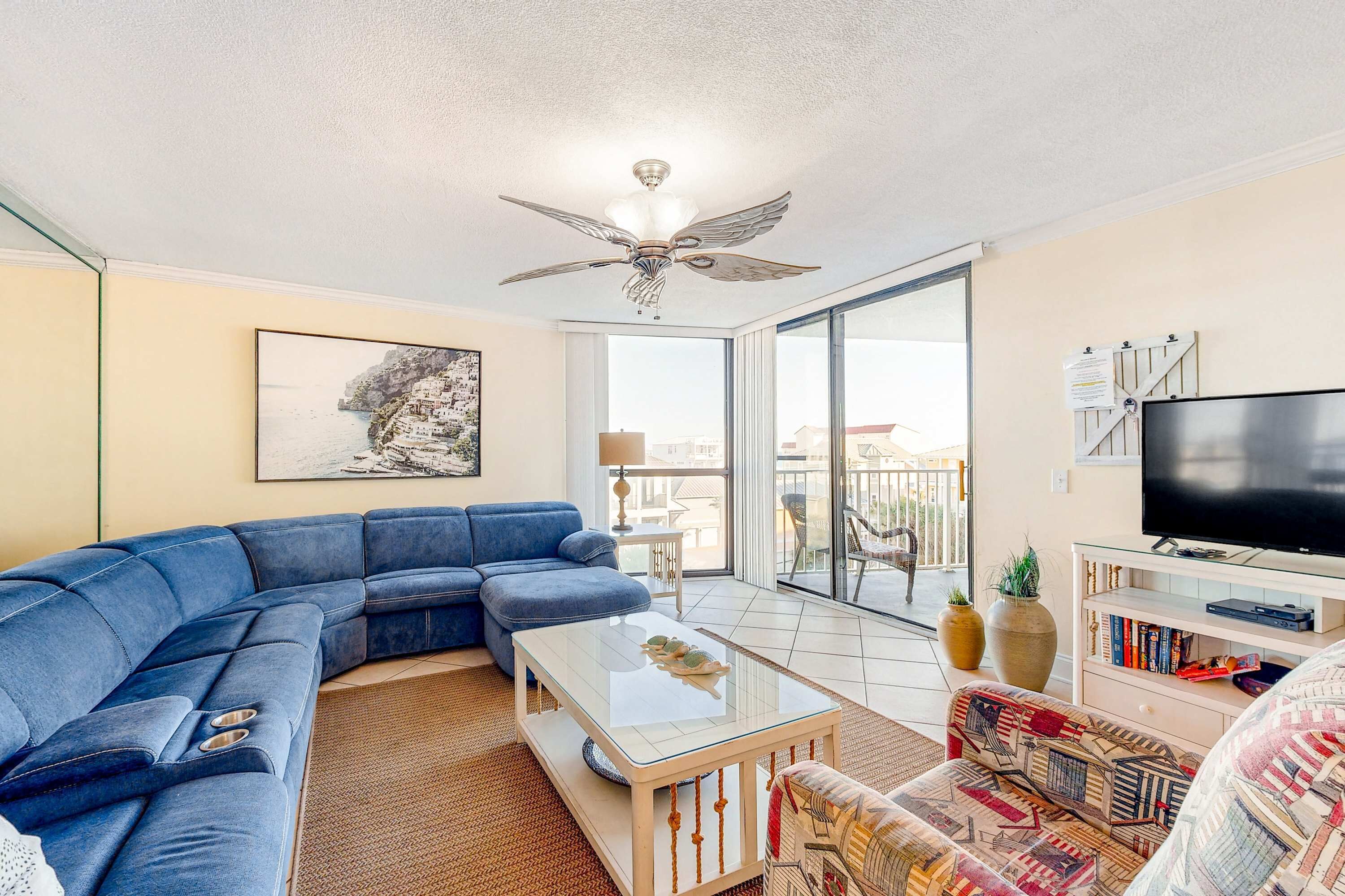 A living room with a sectional sofa, a glass coffee table, a patterned armchair, a flat-screen TV, a ceiling fan, a large window, and a balcony with a view of buildings.