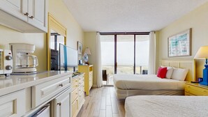 Ett hotellrum med ett litet kök, en säng med vita sängkläder och röda kuddar, ett stort fönster med utsikt och en inramad tavla på väggen.