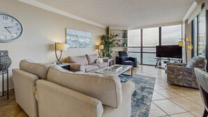 Ein Wohnzimmer mit einer Couch, Sesseln, einem Couchtisch, einem Fernseher und einem großen Fenster mit Meerblick.